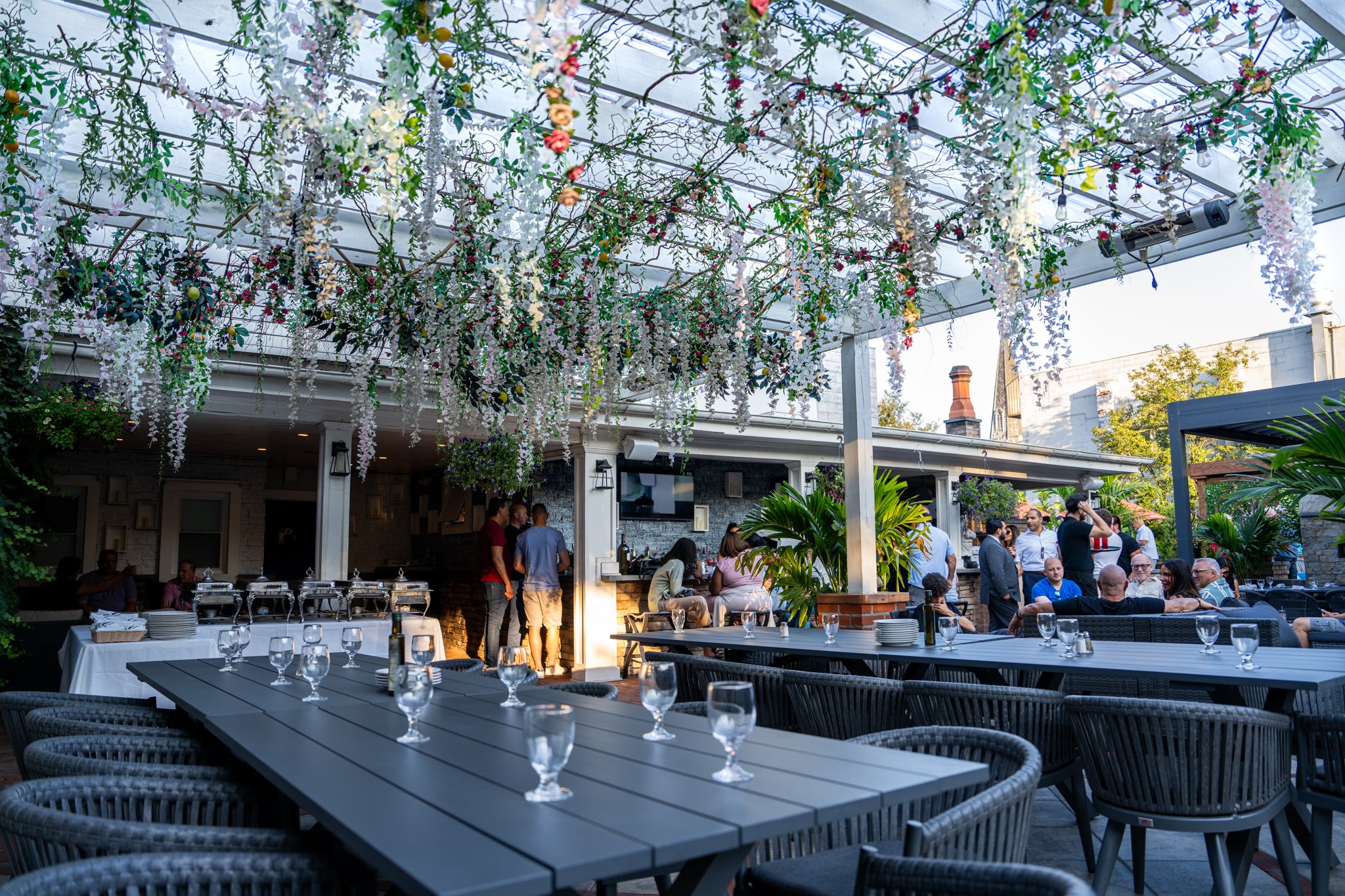 Outdoor dining area decorated with hanging floral arrangements and string lights, with people socializing and seated at tables.