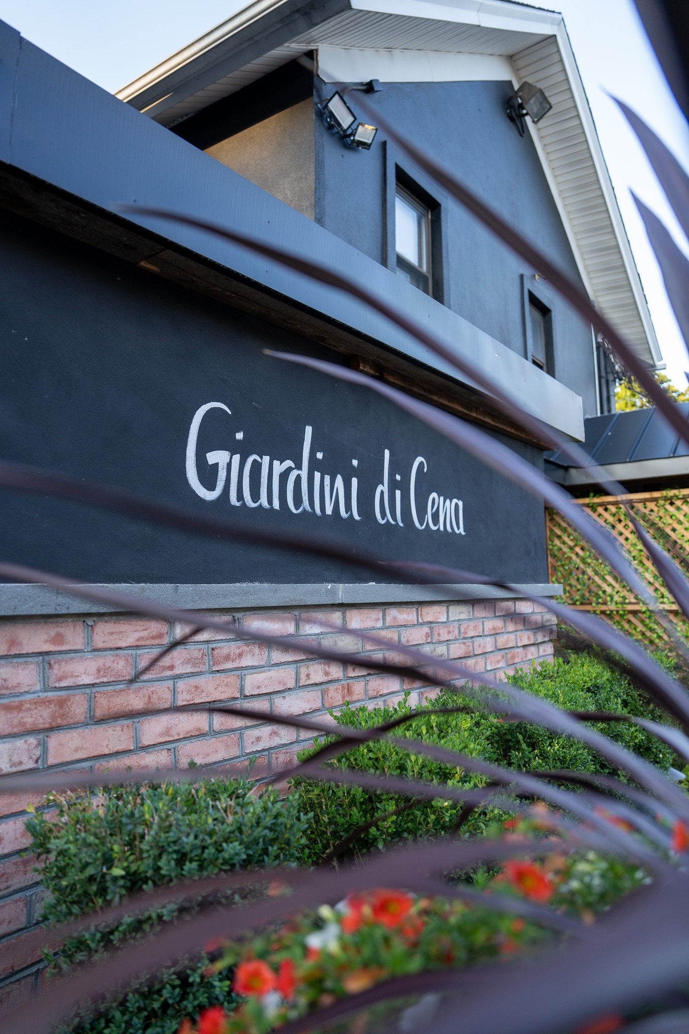 Exterior of a building with a black sign reading 'Giardini di Cena', some greenery and flowers in front, with the upper part of the building visible.