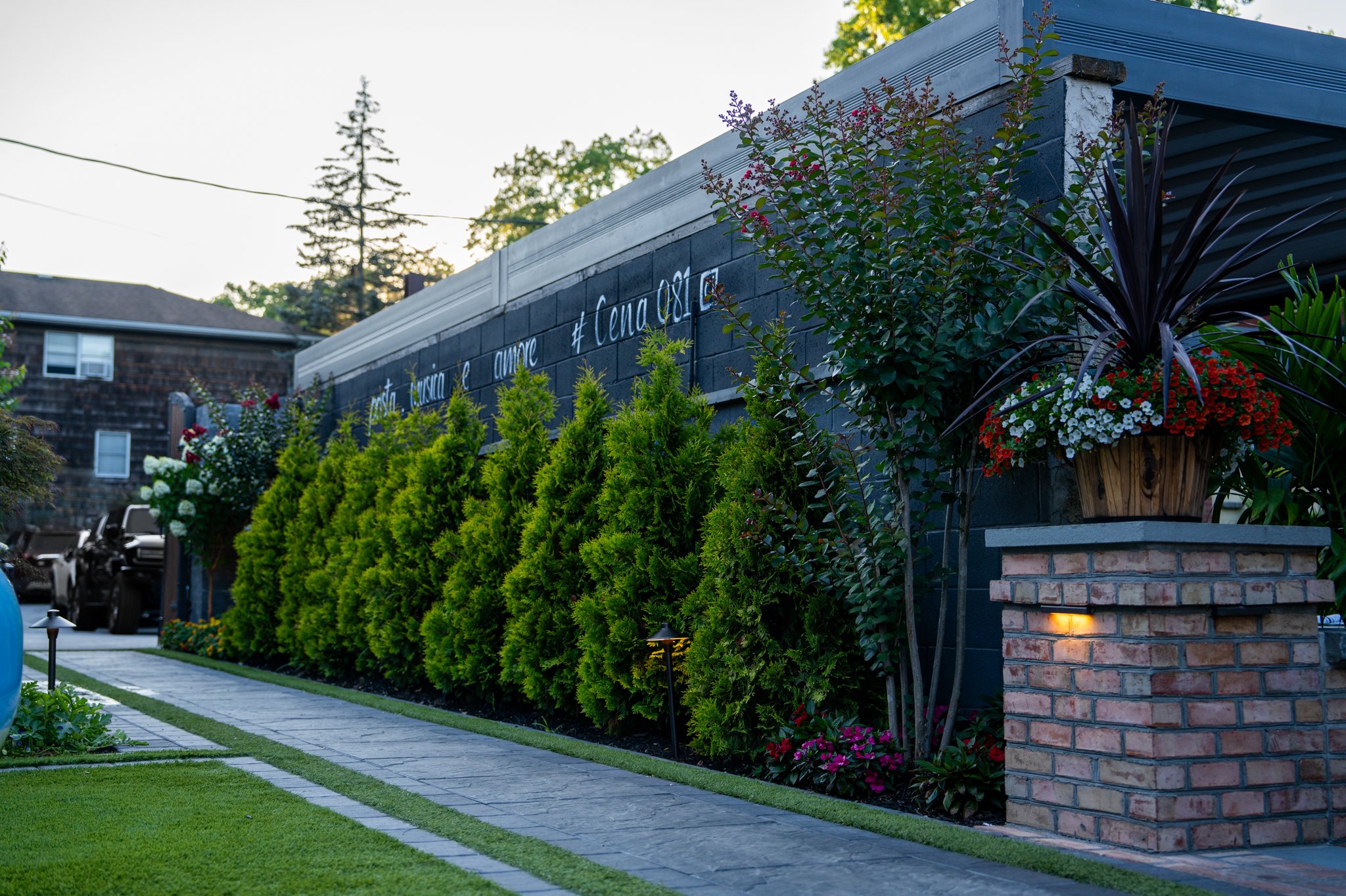 Front yard with green bushes, flowers, and a brick pillar with a flower pot on top, next to a dark brick wall with writing.