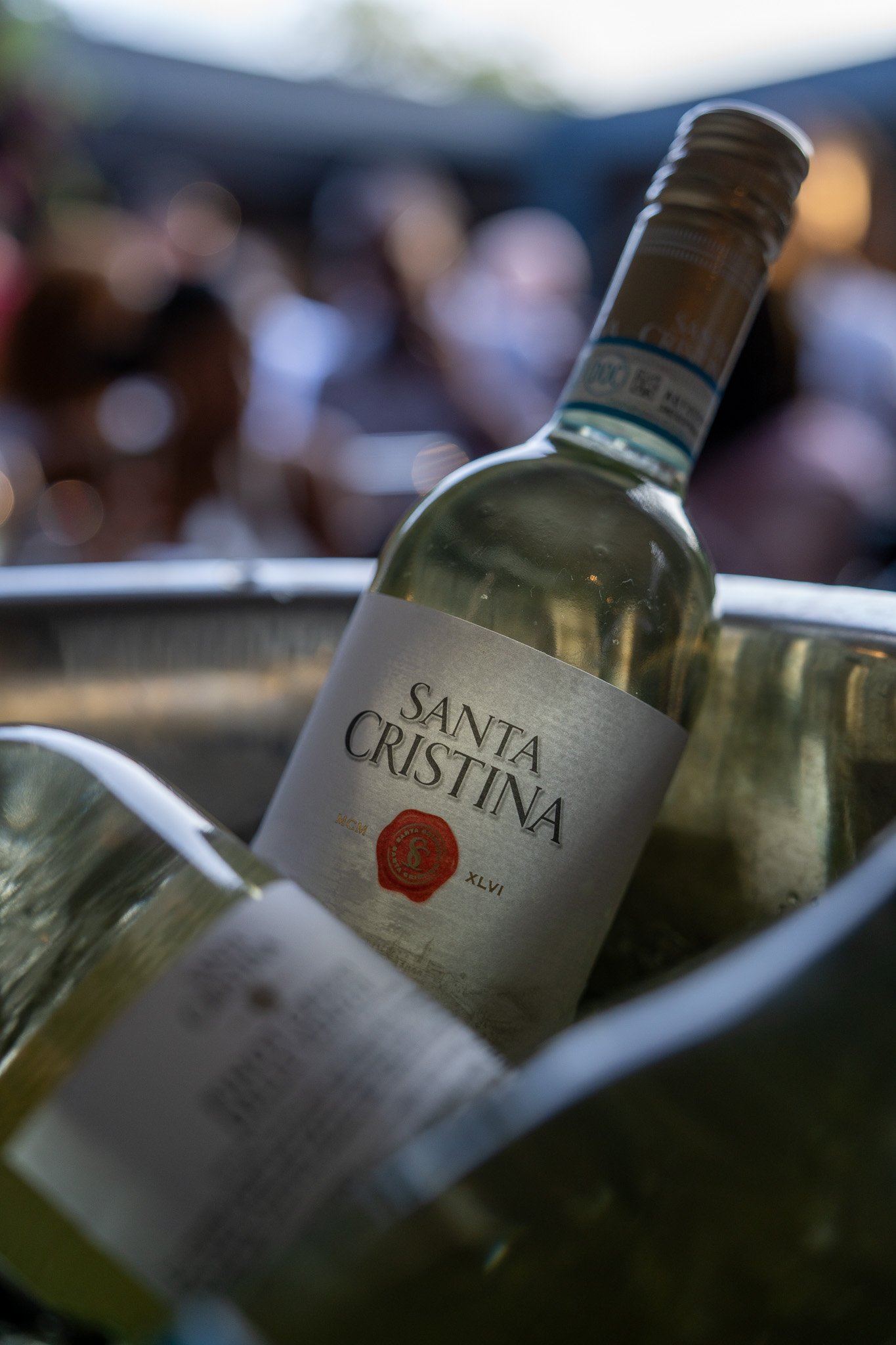 A bottle of Santa Cristina wine chilling in an ice bucket at an outdoor event with blurred people in the background.