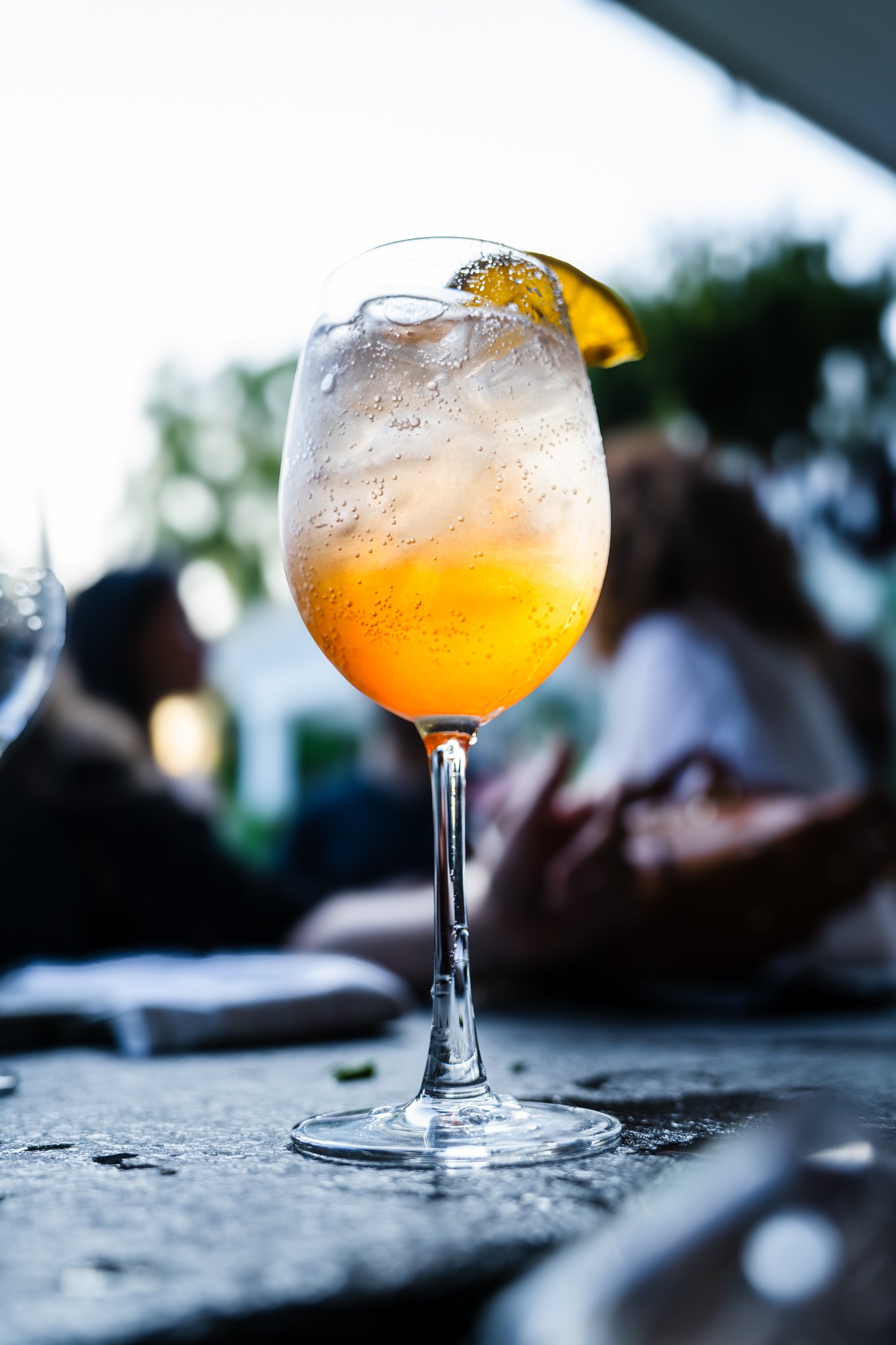 A tall wine glass filled with a bubbly orange-colored cocktail, garnished with a lemon wedge, on a table with blurred people in the background.