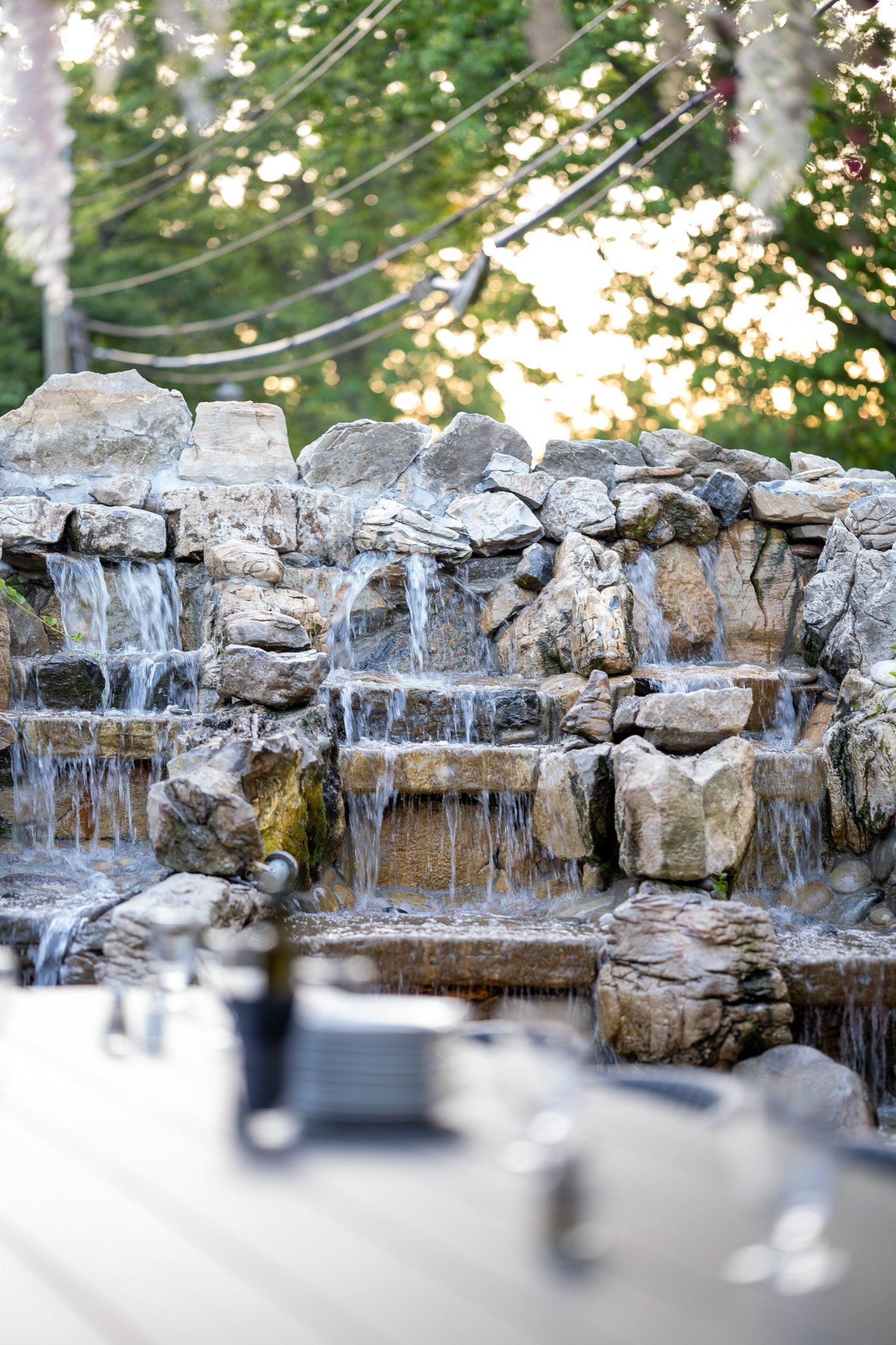A stone water fountain with multiple small waterfalls and rocks, set outdoors with trees and string lights in the background.