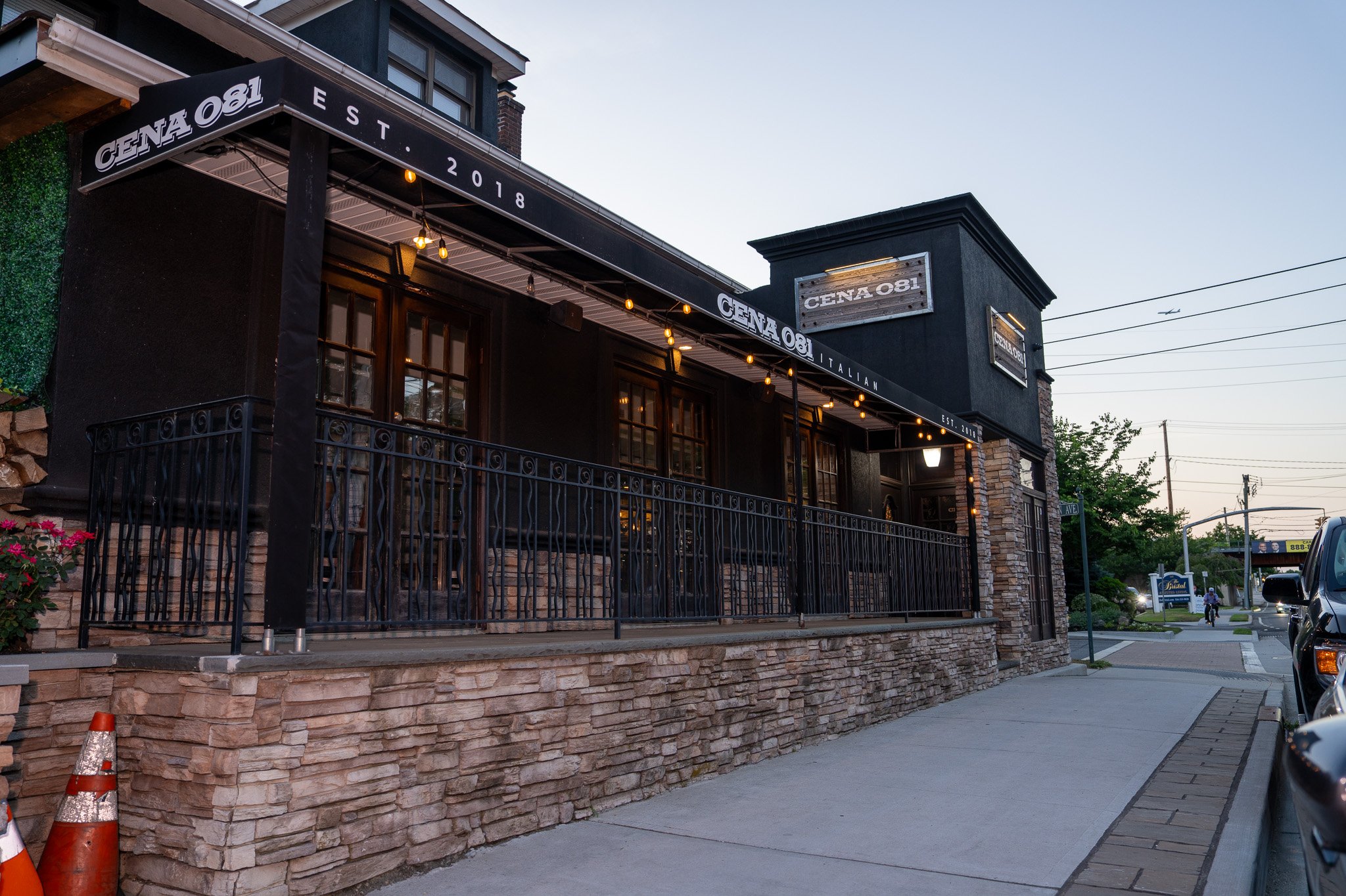 Exterior view of a black-painted Italian restaurant called Cena C81 with a stone foundation, black metal railings, and string lights hanging from the awning, during dusk.