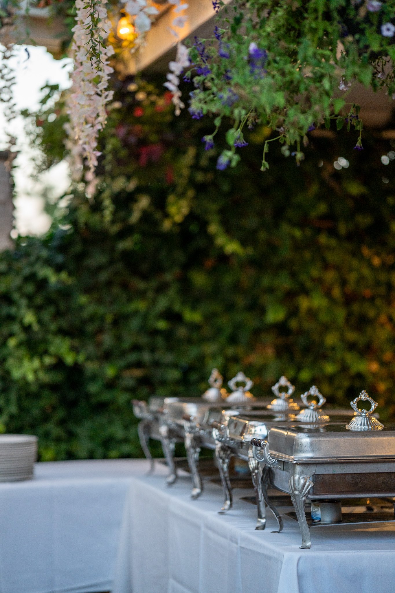 Chafing dishes on a table at an outdoor event, with hanging flowers and greenery in the background.
