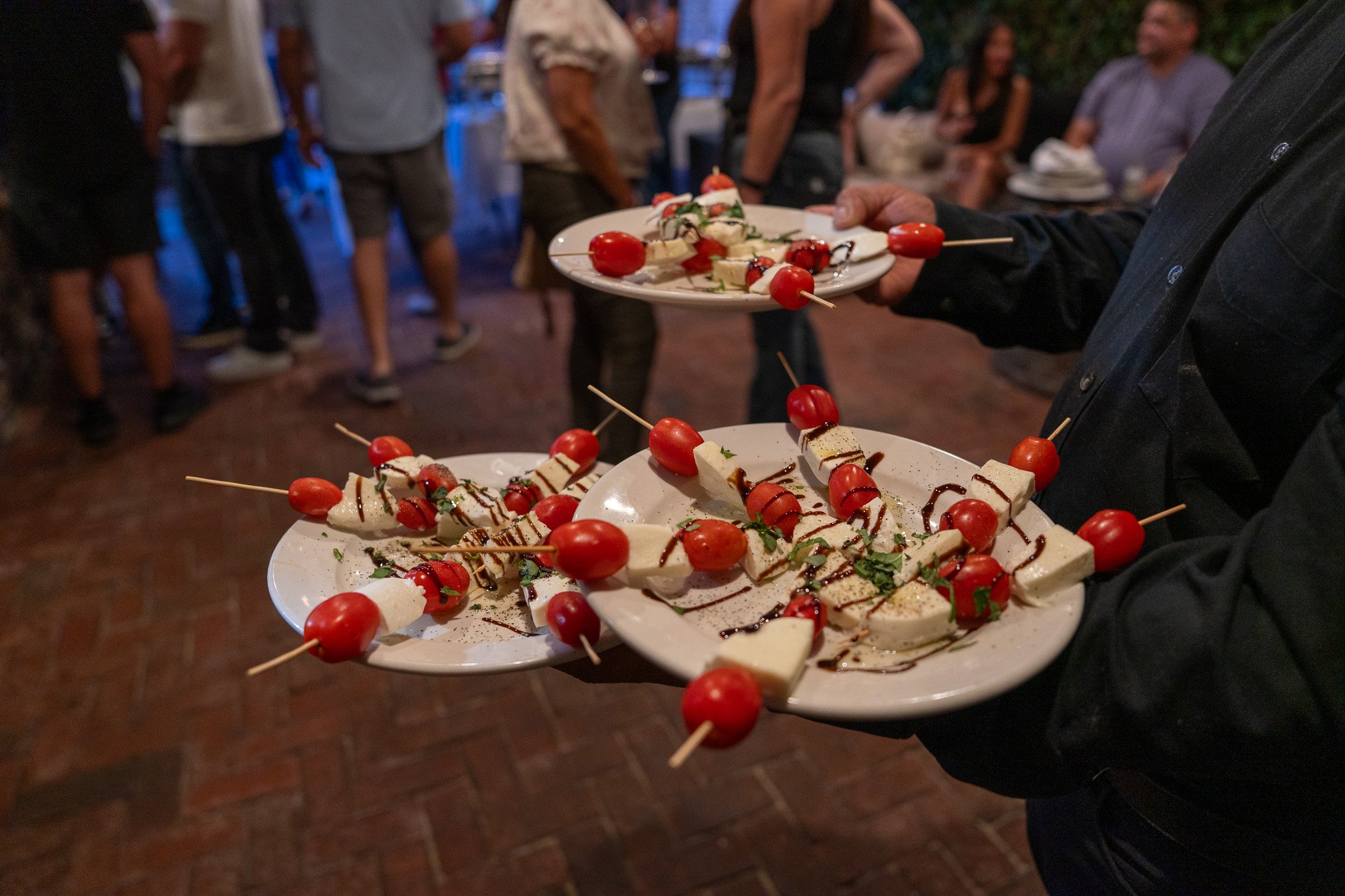 Person holding three plates of caprese skewers with cherry tomatoes, mozzarella, basil leaves, and drizzled balsamic glaze at an outdoor gathering.