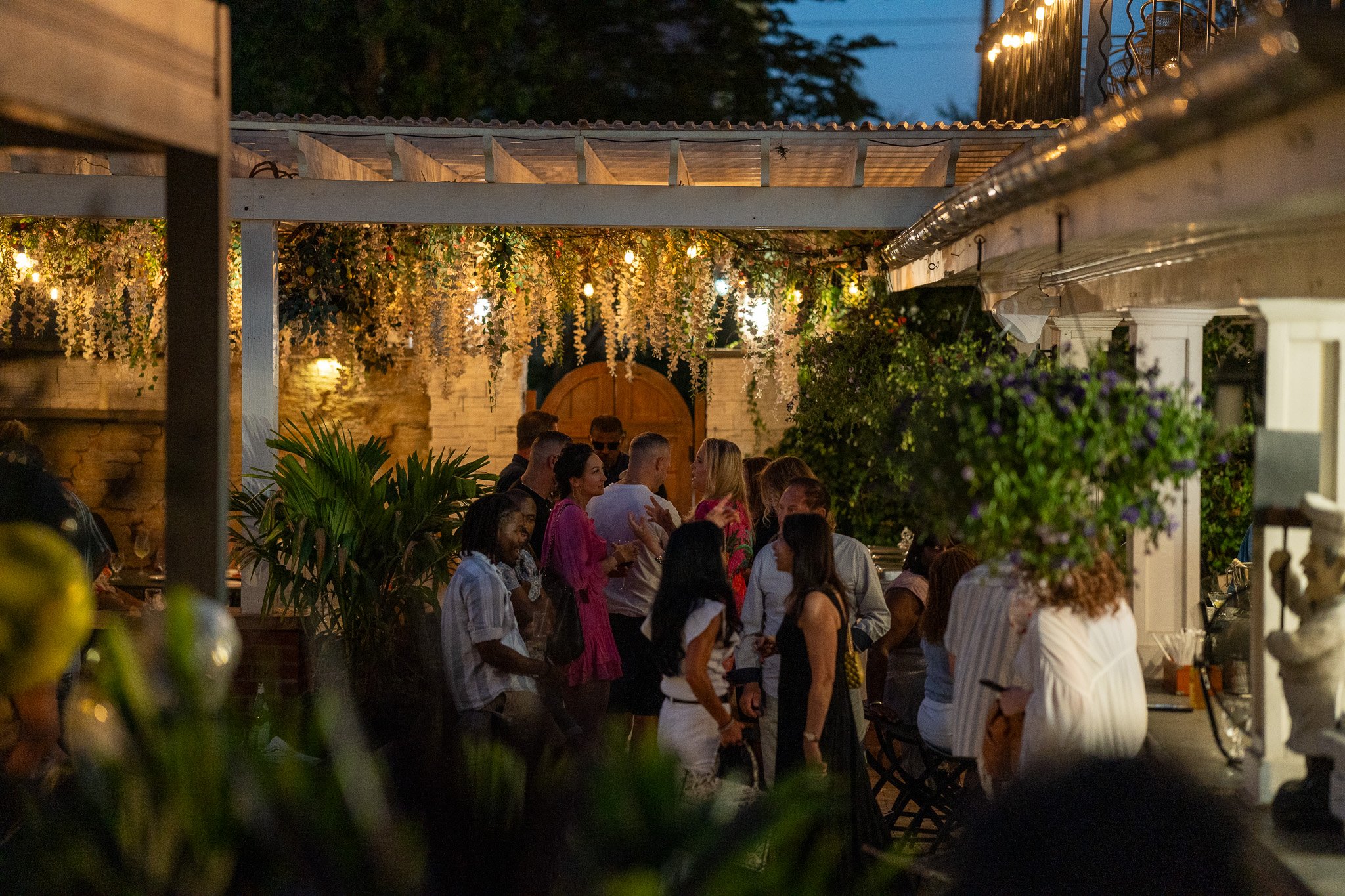 People socializing at an outdoor evening gathering under string lights and hanging floral decorations.