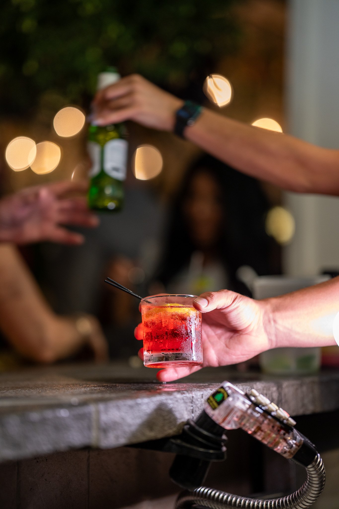 Person holding a red cocktail glass with a black straw at a bar counter, other people in the background with blurred lights, one person reaching with a bottle, and a hookah hose on the counter.