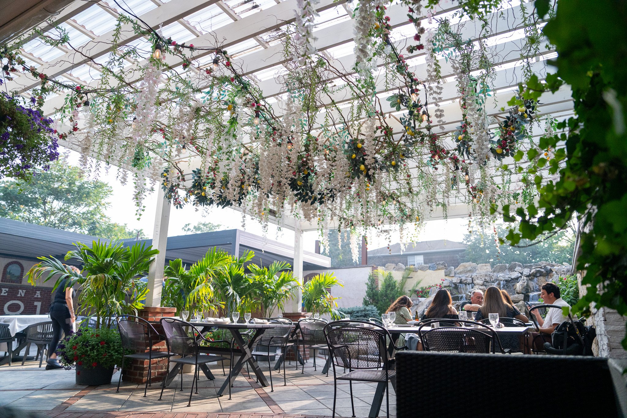 Outdoor patio with hanging floral arrangements on a pergola, lush green plants, and a group of people dining at tables with wine glasses.