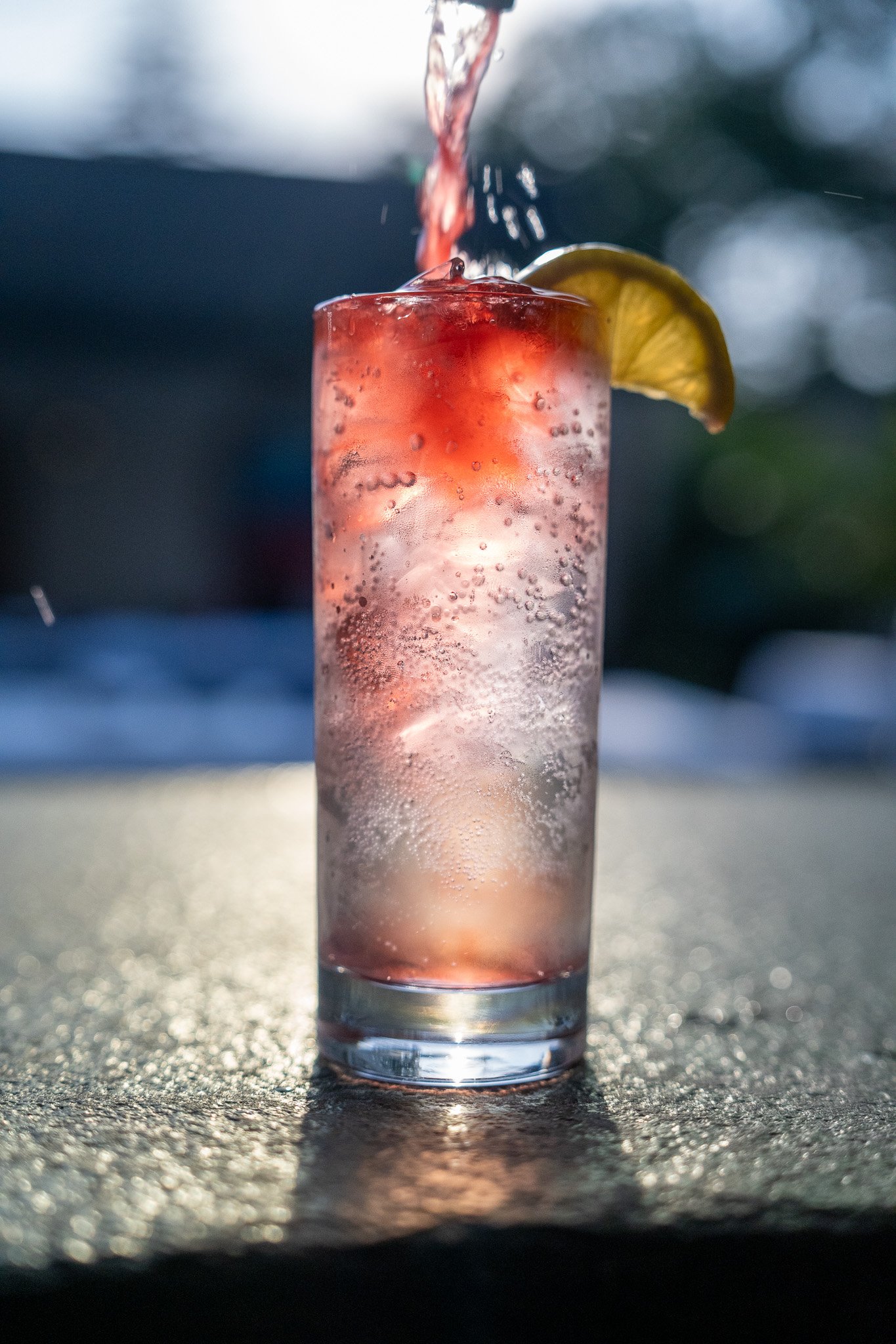 A tall glass of a pinkish fizzy drink with ice and a lemon wedge on the rim, being poured with water.
