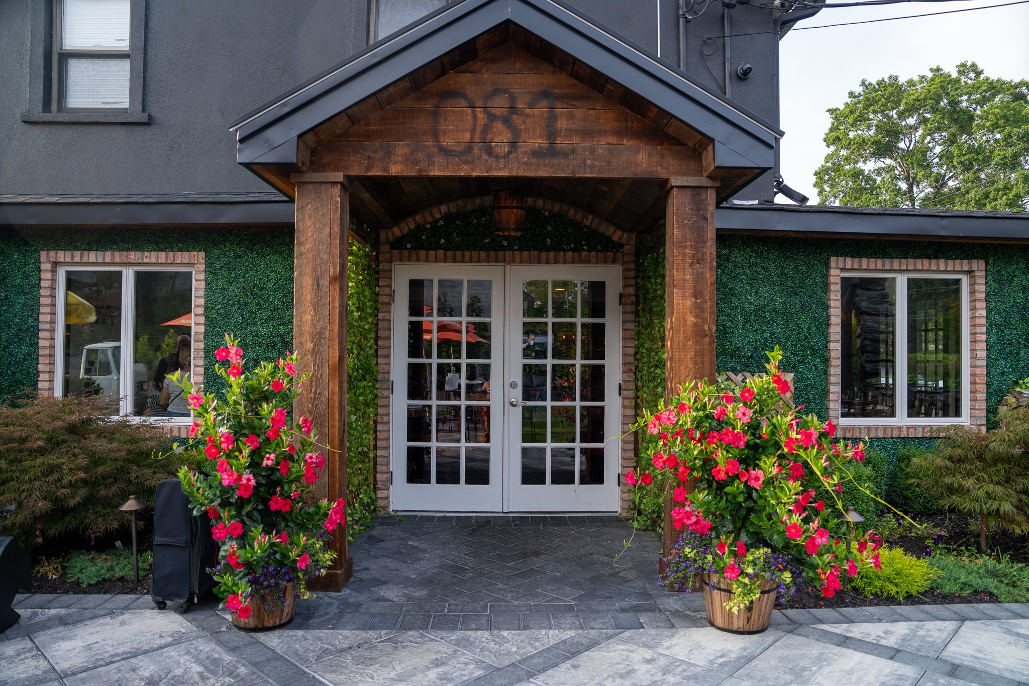 Entrance of a building with a wooden portico, double glass doors, and two large flower pots with pink flowers on each side, surrounded by greenery.