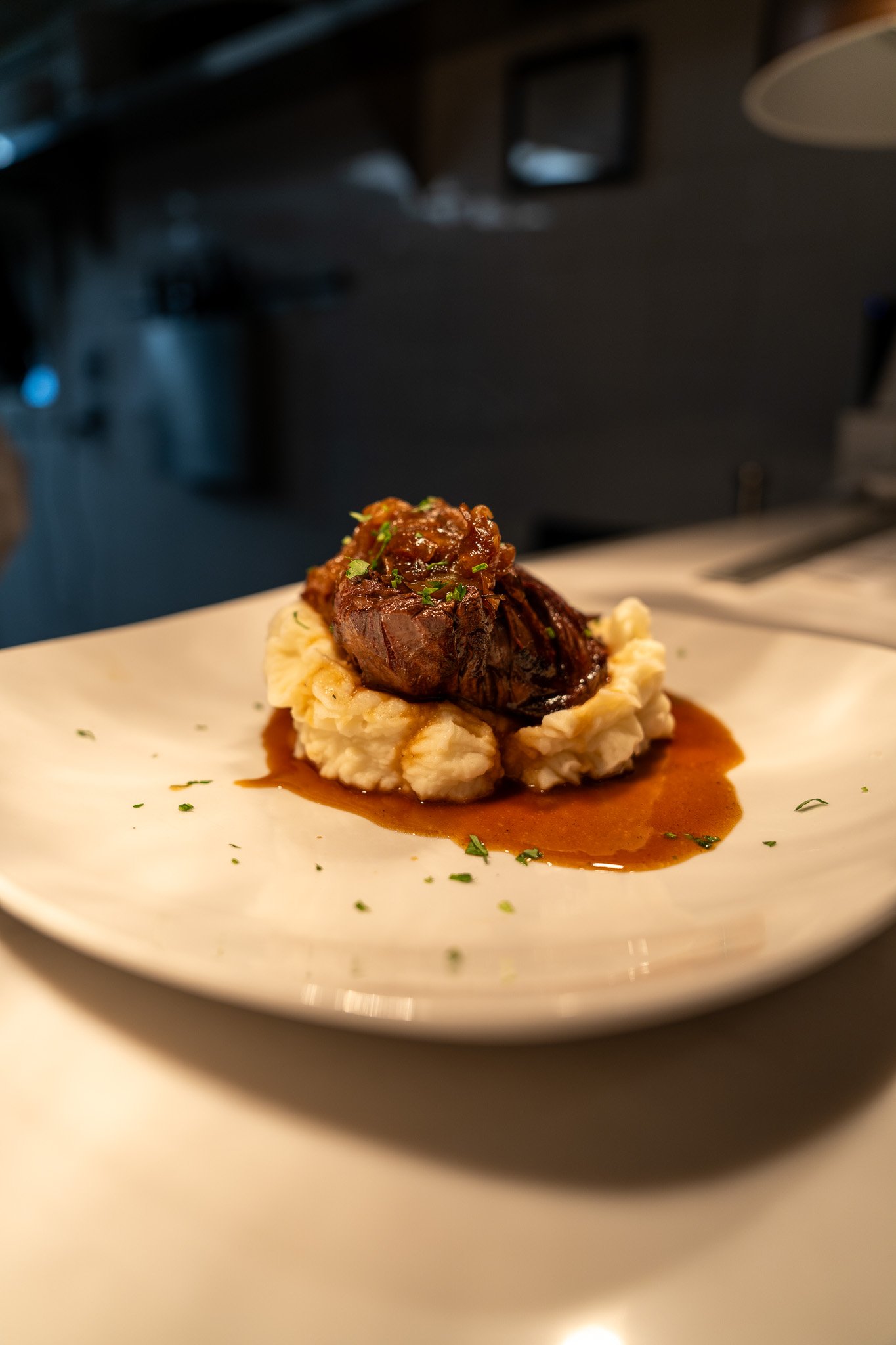 A plated dish of braised beef short rib served over creamy mashed potatoes with gravy, garnished with chopped herbs.