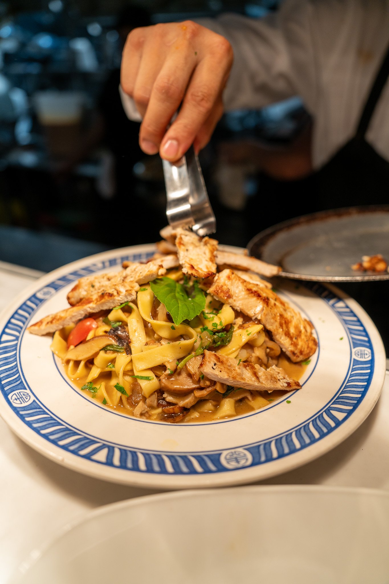 Person placing grilled chicken slices on pasta with mushrooms, herbs, and vegetables.