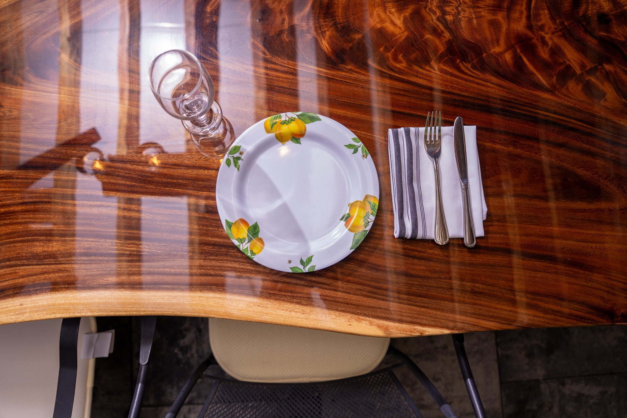 An empty white plate with yellow lemon print border on a wooden table, with a stack of striped napkin, a fork, and a knife wrapped in the napkin, and a clear glass with a curved stem.