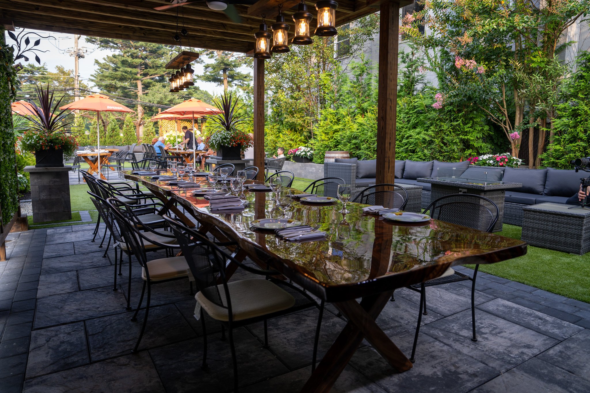 Outdoor restaurant patio with a large polished wooden dining table set with plates, silverware, and glasses, surrounded by black chairs, and an outdoor seating area with a sectional sofa. There are potted plants and flowers, trees, and umbrellas in t