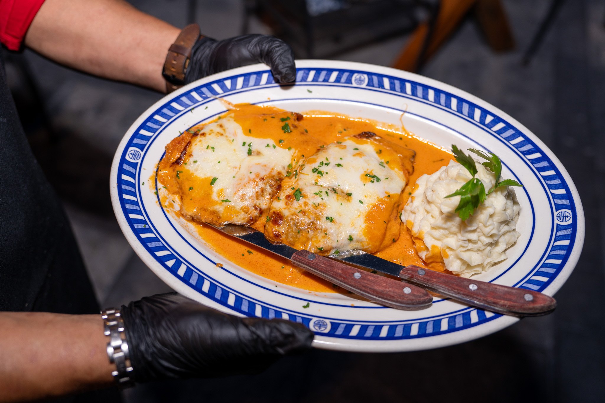 A person wearing black gloves and a watch holds a decorative blue and white oval plate with two baked dishes topped with melted cheese, garnished with chopped herbs, and a side of mashed potatoes with parsley.