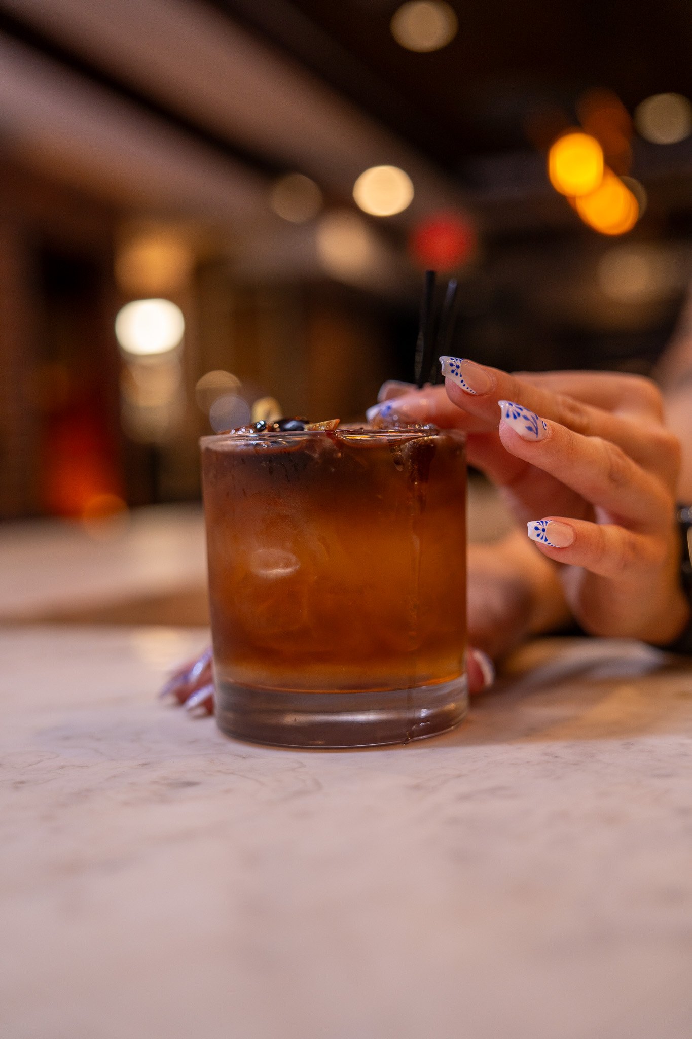 Person with decorated nails reaching for a glass of iced cocktail in a dimly lit bar or restaurant.