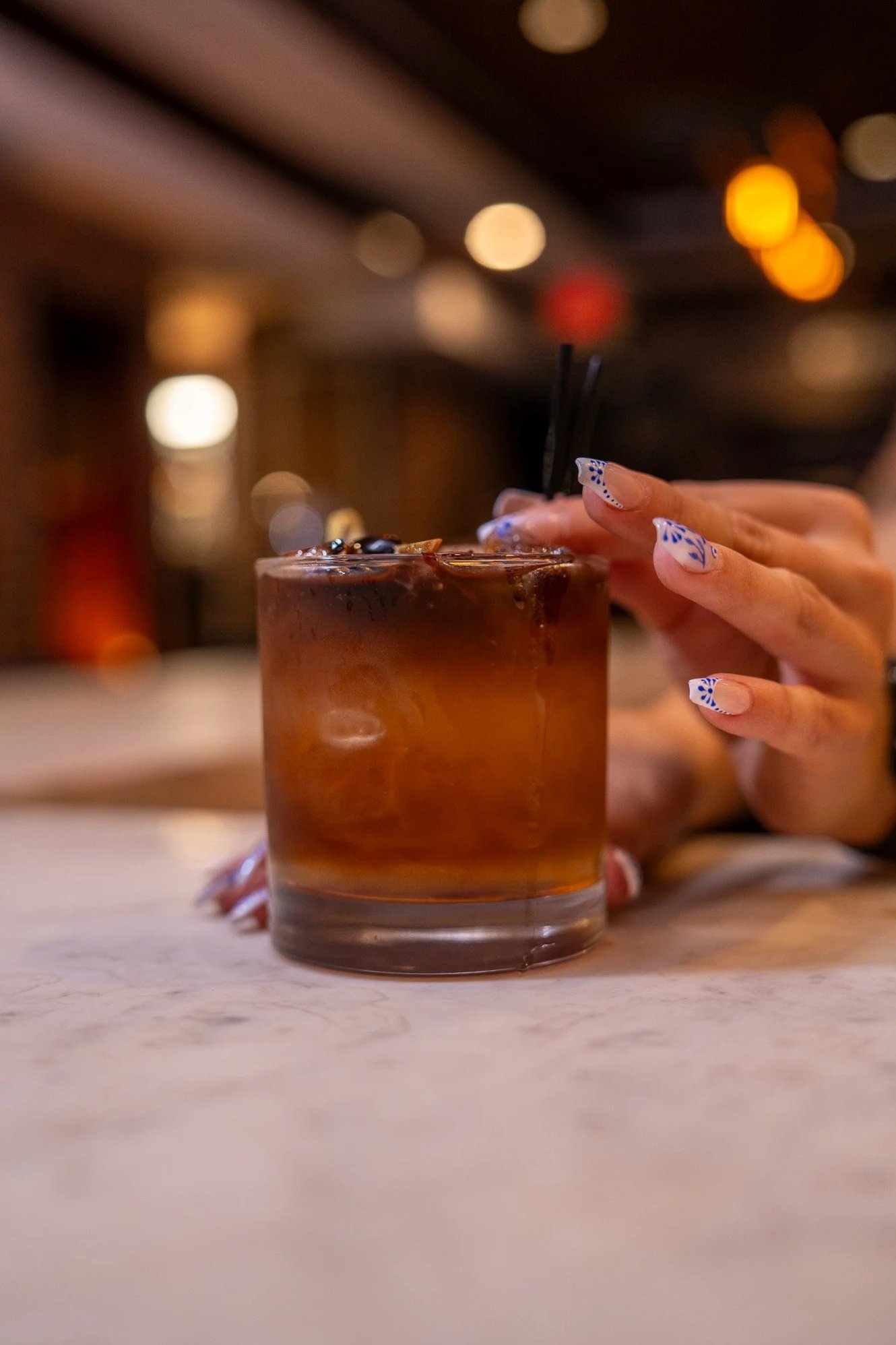 A person’s hand with decorated nails holding a glass of iced coffee or cocktail with a dark liquid and ice, on a marble countertop in a dimly lit bar or restaurant setting.