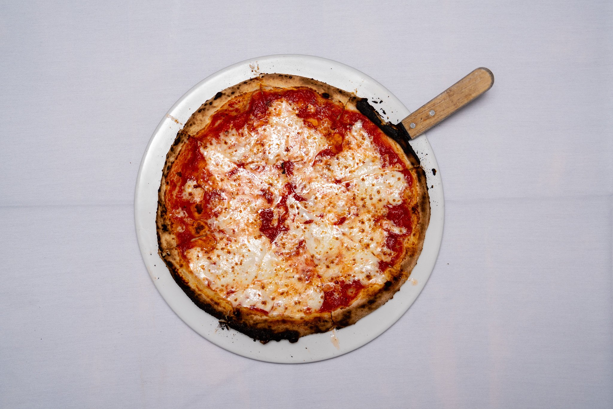 A cheese pizza on a white plate with a metal pizza cutter resting on the side.