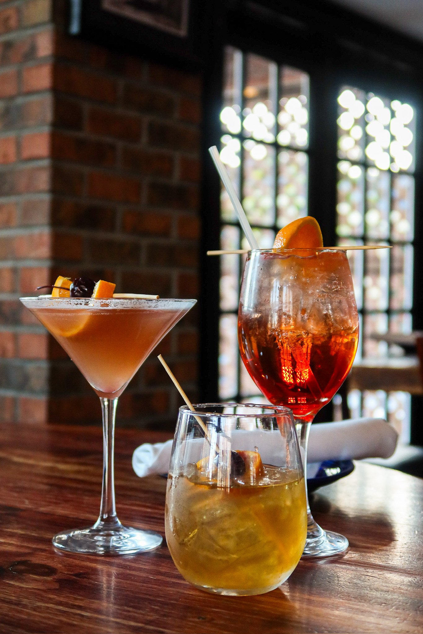 Three cocktails on a wood bar counter with a brick wall and window in the background. One appears to be an orange-colored drink with ice and a slice of orange, another is a brownish drink with garnishes, and the third is a yellowish drink with ice.