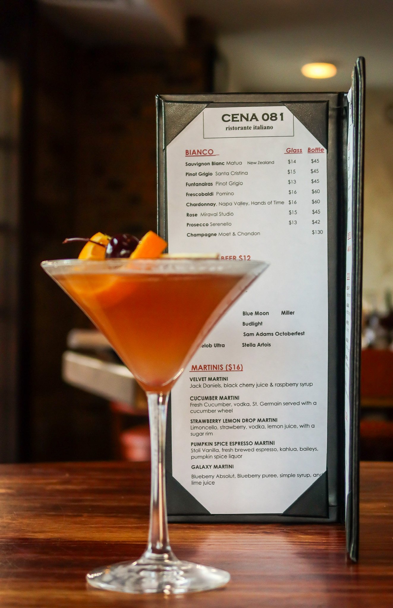 A cocktail in a martini glass garnished with orange slices and a cherry, placed in front of a restaurant menu on a wooden table.