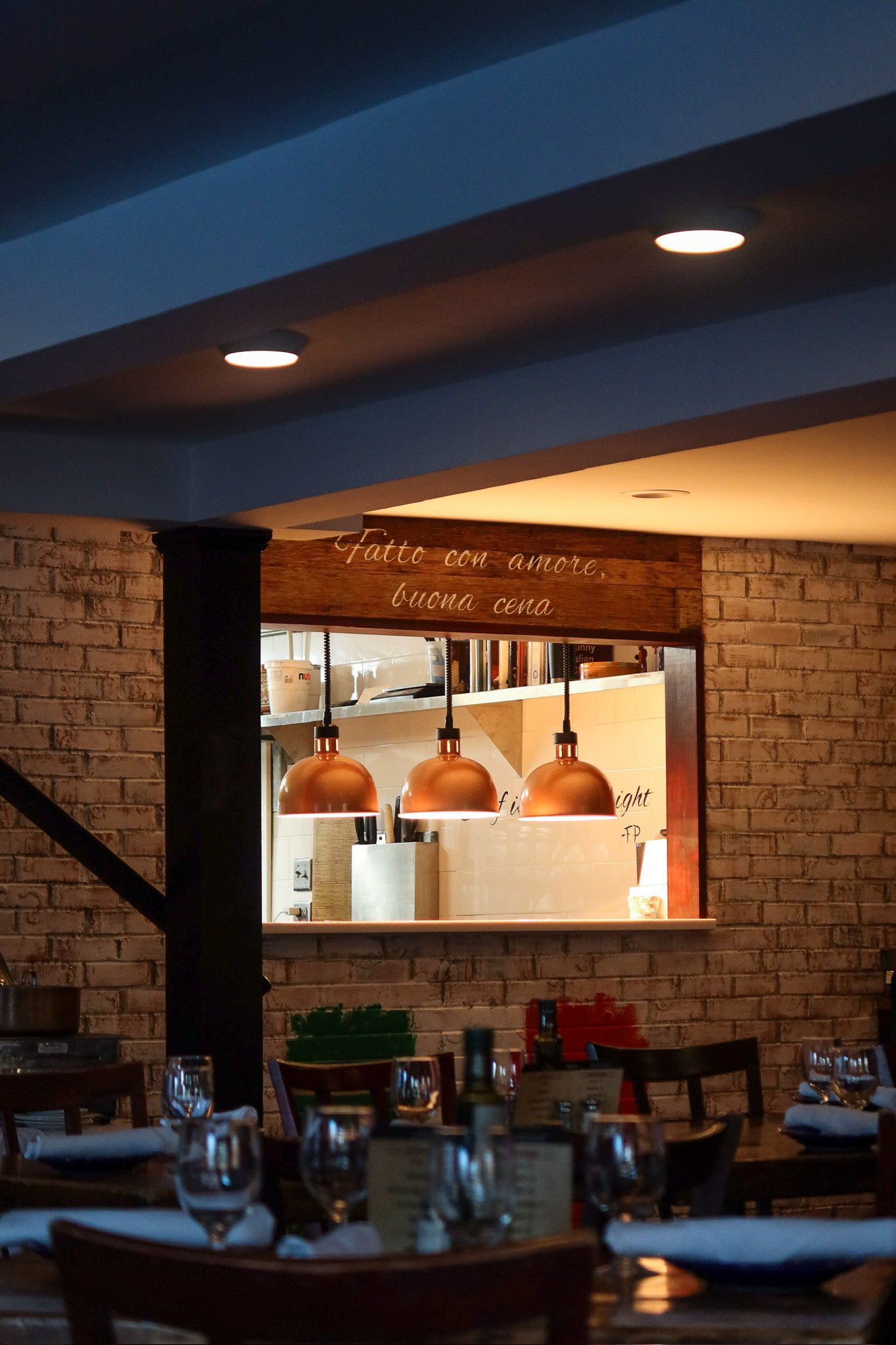 Interior of a restaurant with tables set with glasses and napkins, warm lighting, exposed brick walls, and a view into the kitchen through a window with a wooden sign above that reads in Italian 'Fatto con amore, buona cena'.