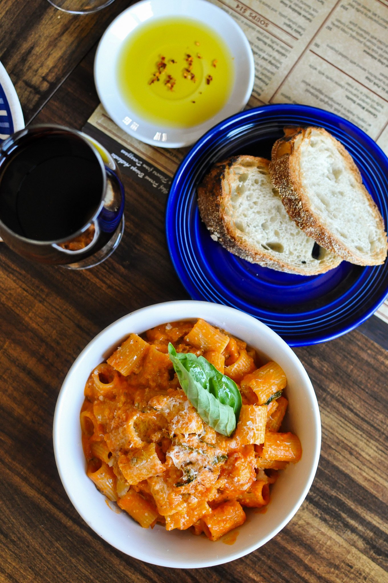 A meal on a wooden table including a bowl of pasta with tomato sauce and cheese, slices of bread on a blue plate, a small bowl of olive oil with chili flakes, and a glass of red wine.