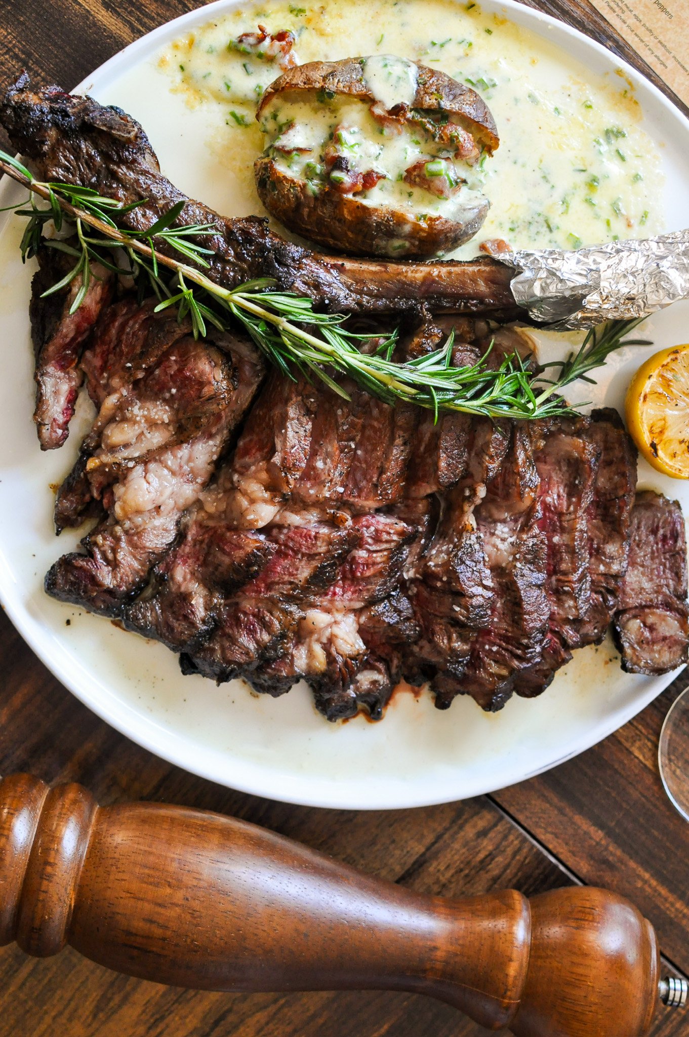 A plate of grilled steaks garnished with rosemary, with a twice-baked potato topped with cheese and bacon, a halved lemon, and a sauce with herbs on a wooden table.