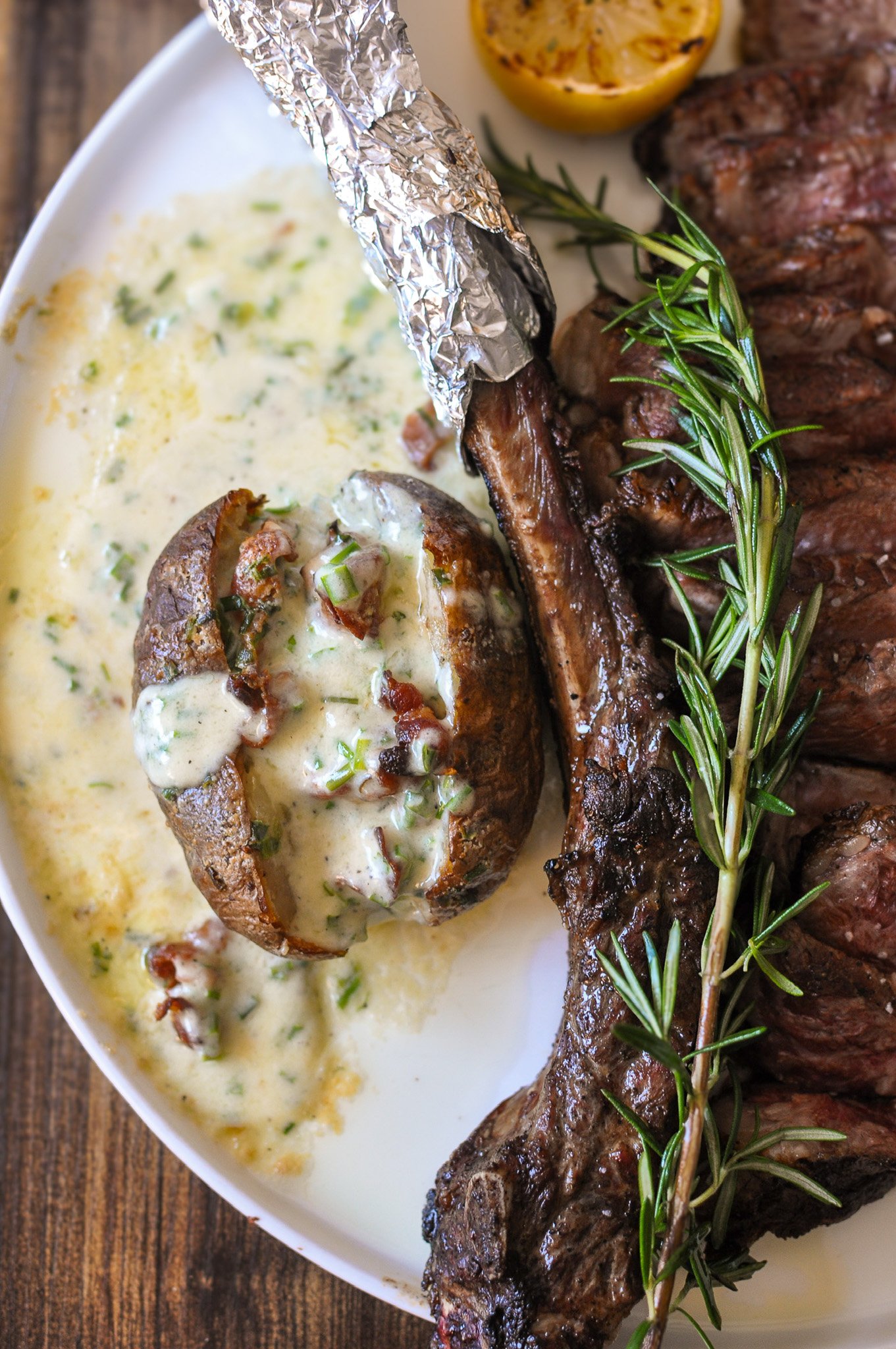 A plate of cooked steak garnished with a sprig of rosemary, a baked potato topped with sour cream and bacon bits, and a small dish of grilled lemon.