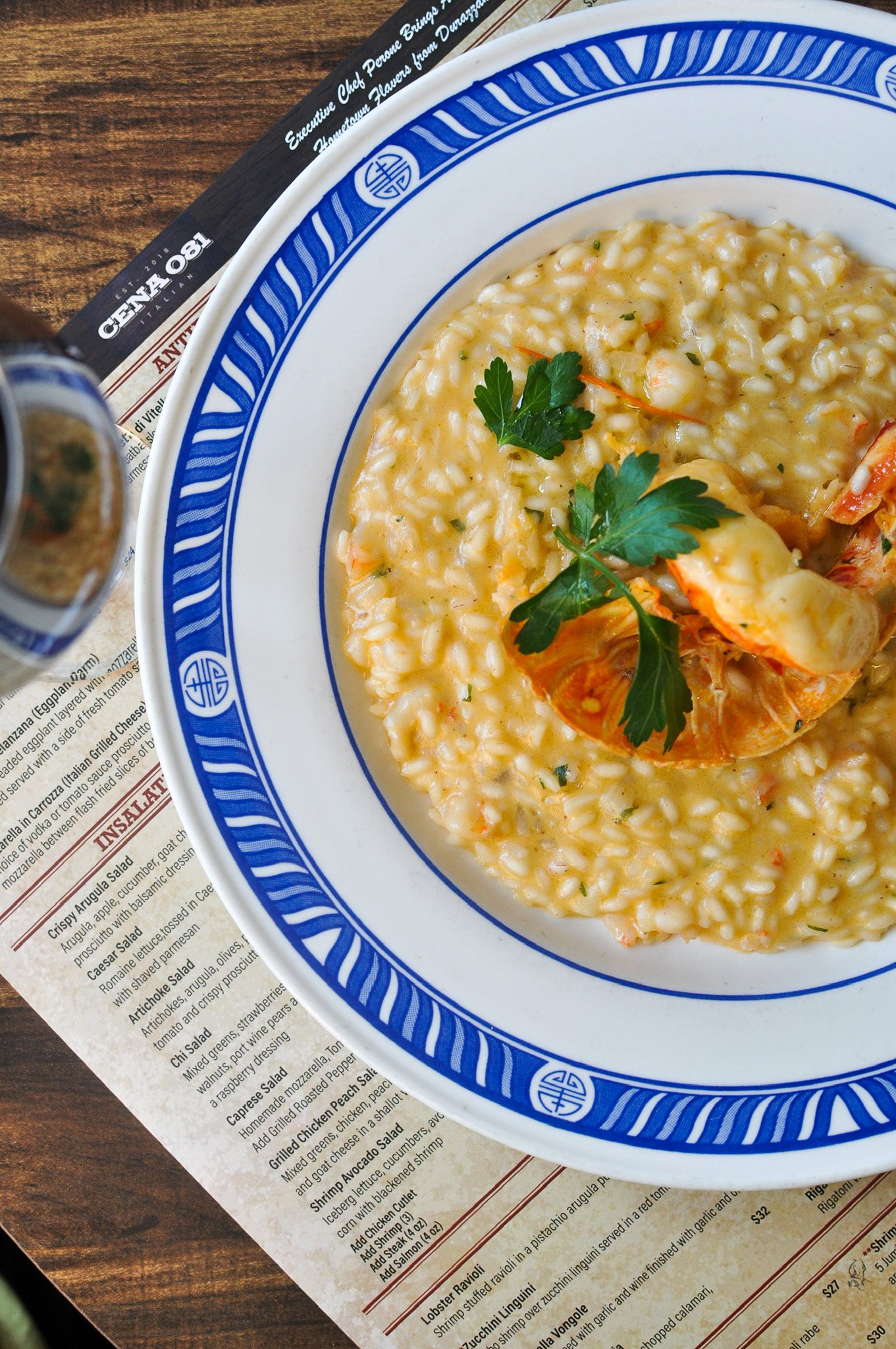 A plate of lobster risotto garnished with parsley, on a blue and white patterned dish, placed on a wooden table with a menu partially visible underneath.